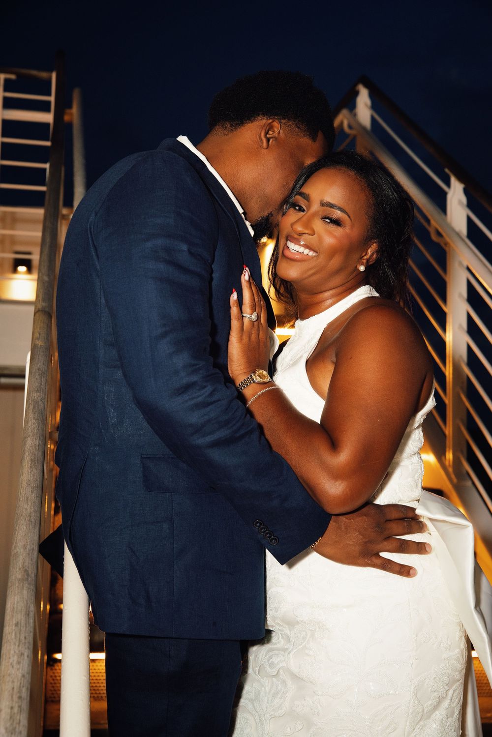 Couple in wedding attire embracing on stairs at night, bride in white dress smiling joyfully at groom in navy suit.