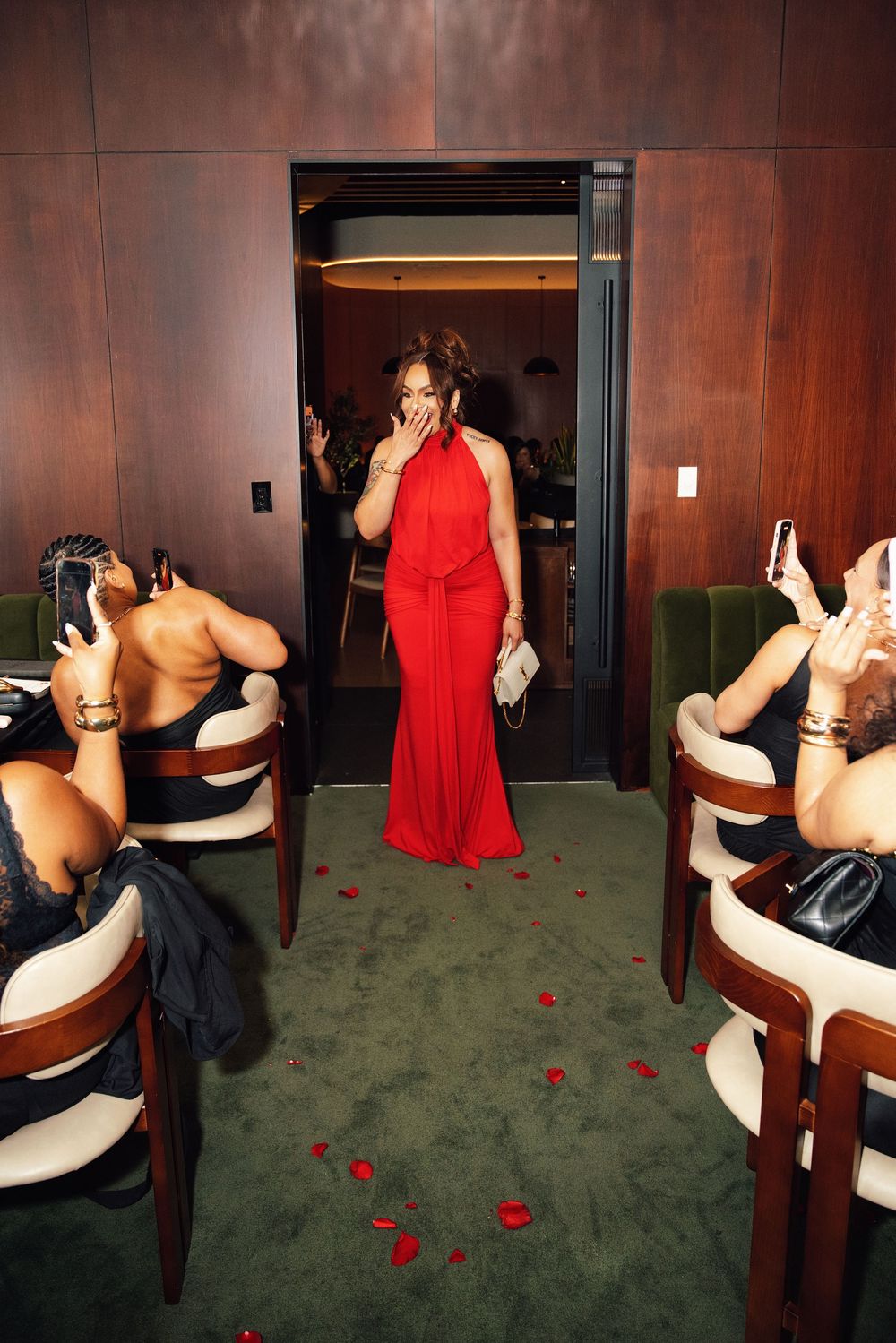 Woman in elegant red gown enters a room as seated guests photograph her, with rose petals on green carpet.