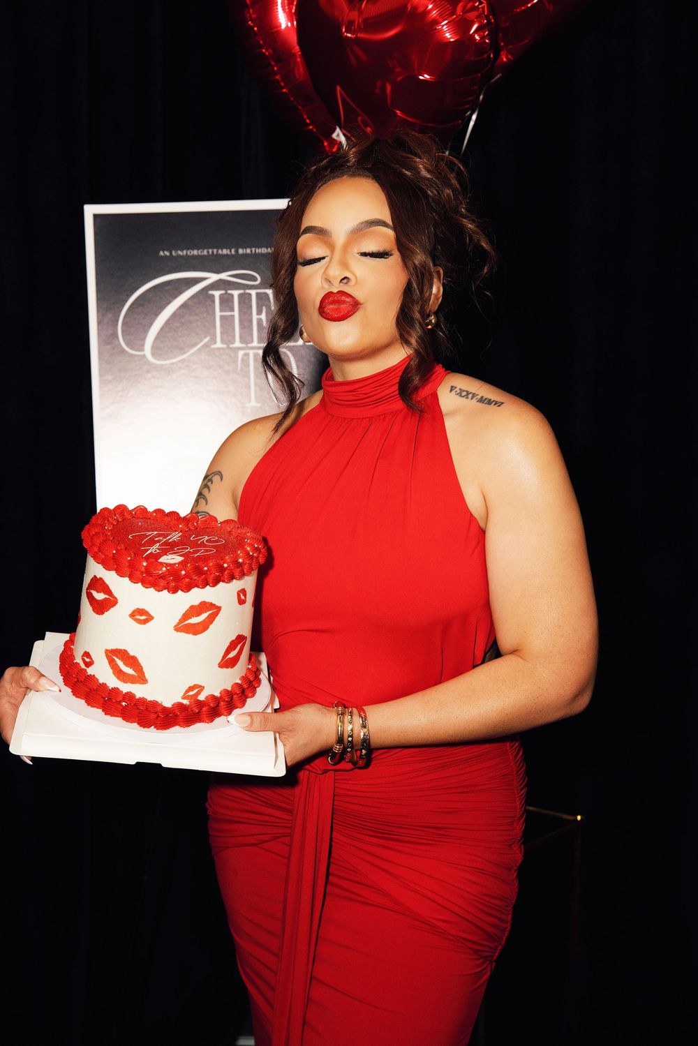 Woman in red dress holding decorated birthday cake with red lips design, red heart balloons in background.