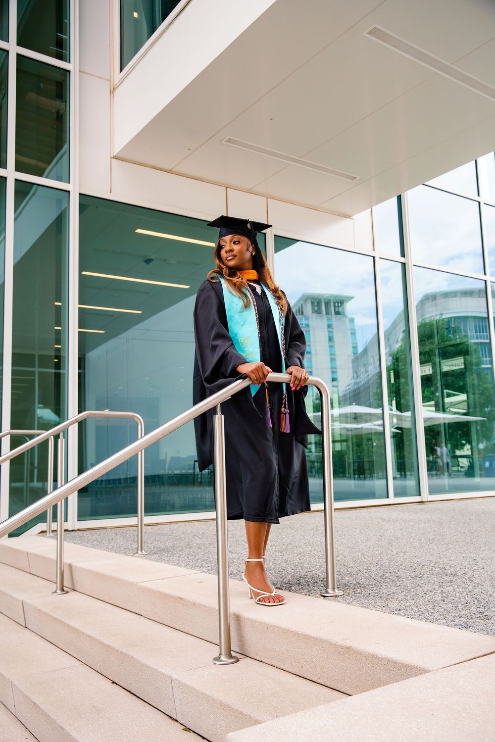 A smiling graduate in black cap and gown with teal stole poses on steps outside a modern glass building.