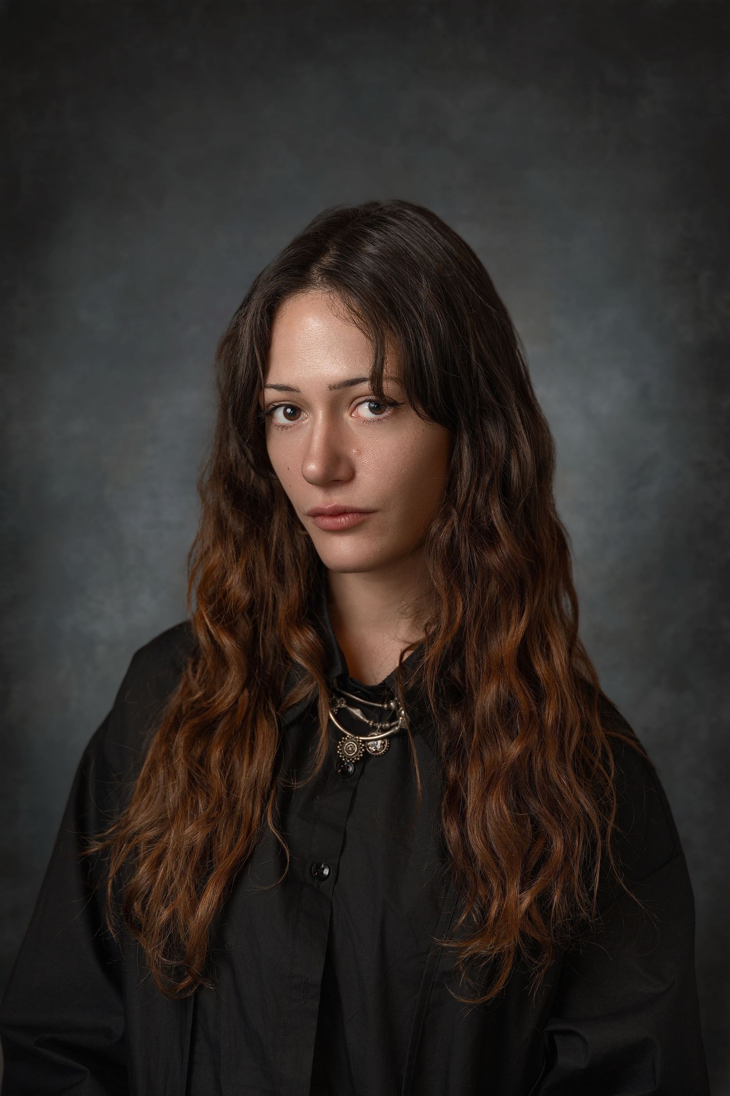 Young woman with long wavy brown hair wearing a black top and gold necklace against a dark gray background.