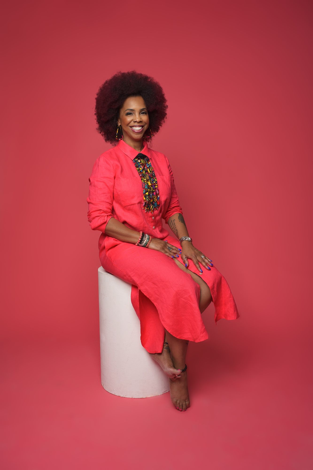 Smiling woman with afro hair wearing a red dress sits on a white pedestal against a red background.