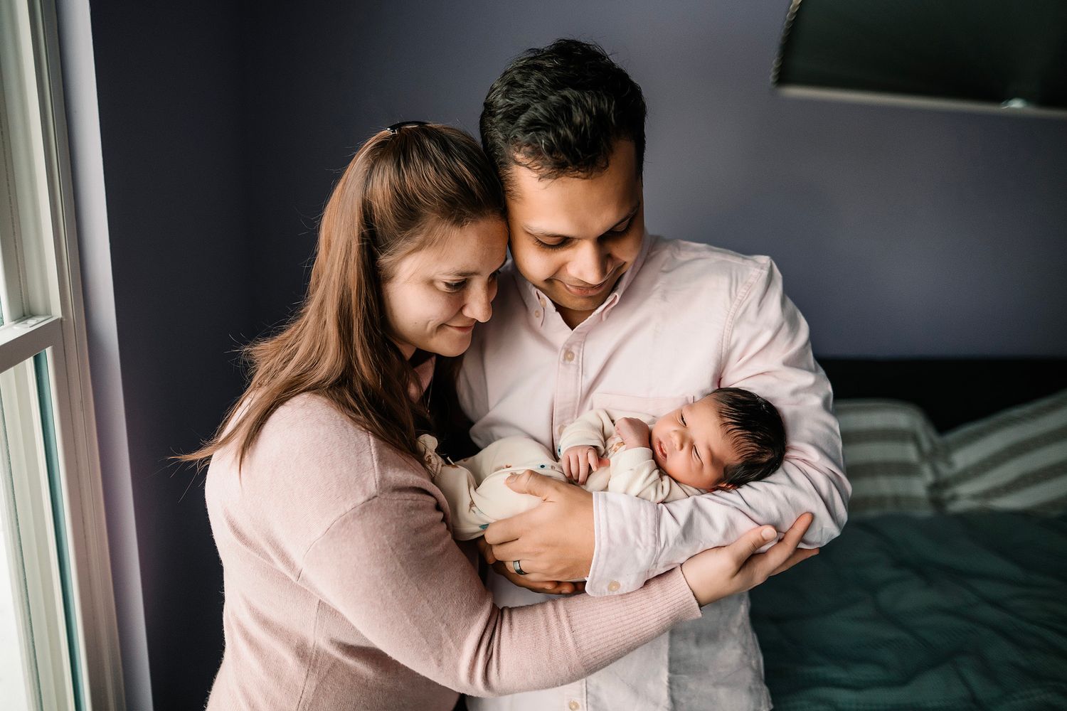 Happy new parents smiling and cradling their sleeping newborn baby together in a cozy bedroom.