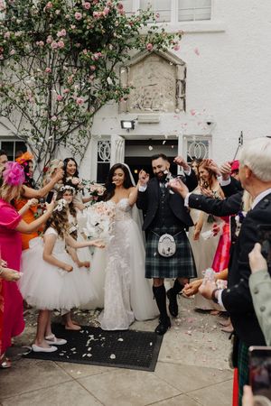 Newlyweds celebrating with confetti shower outside a white venue surrounded by colorful guests.