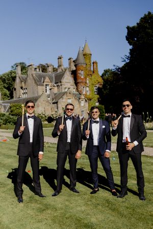 Four groomsmen in tuxedos posing with bow ties on a sunny lawn before a Scottish castle.