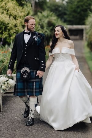 Smiling bride in off-shoulder gown walks hand-in-hand with groom wearing traditional Scottish kilt.