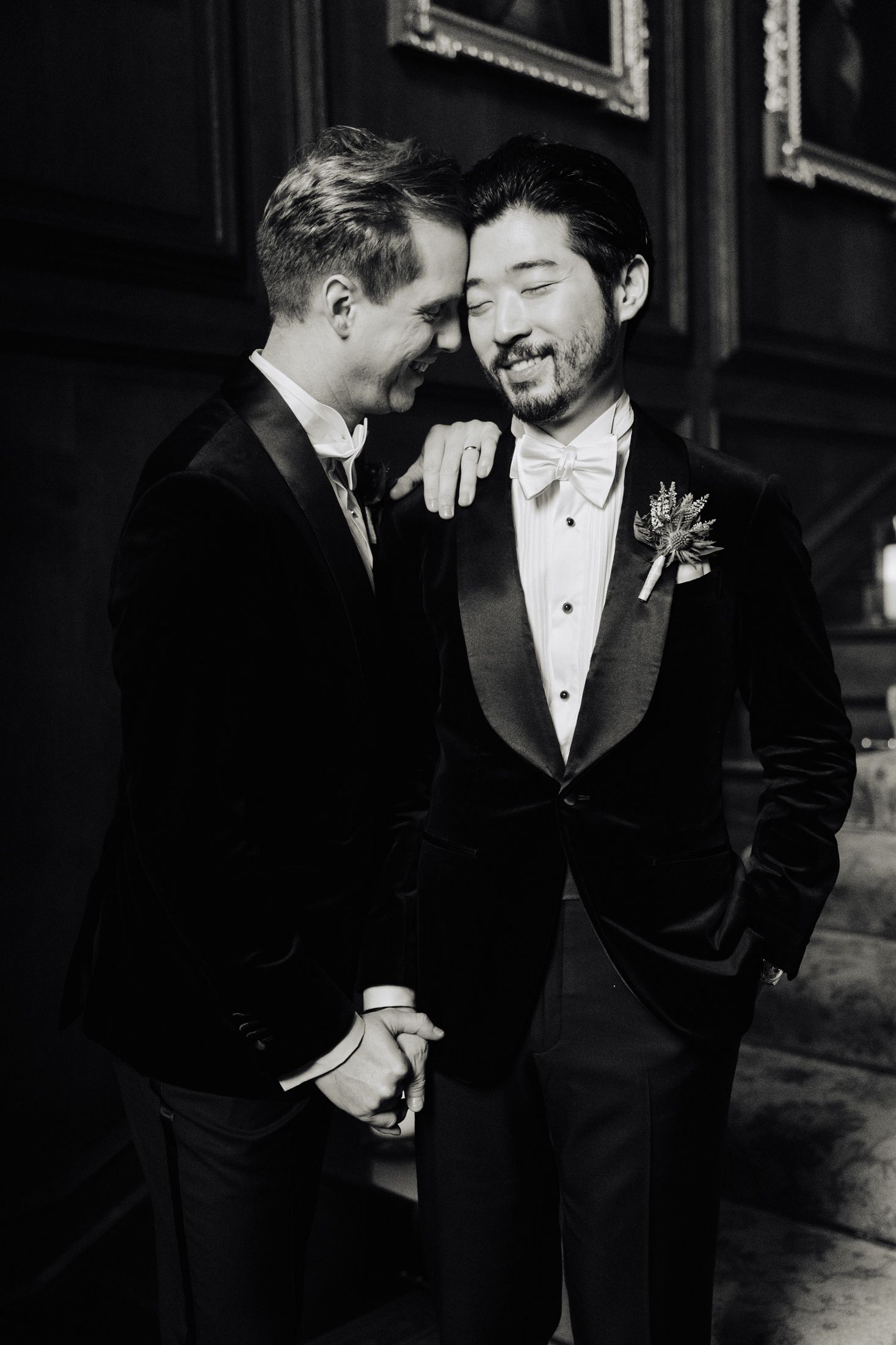 Black and white photo of two grooms in velvet suits sharing a tender moment indoors.