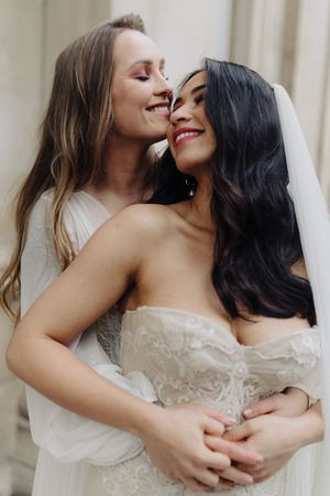 Two brides in white wedding attire share an intimate moment, smiling and embracing each other.