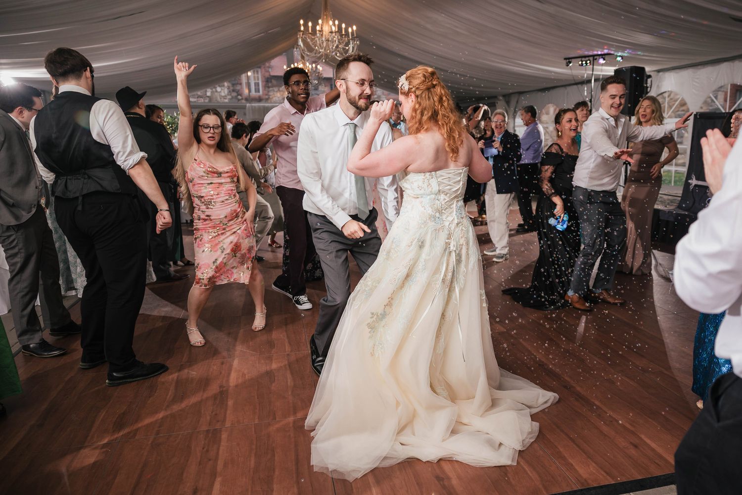 Wide view of the bride and groom dancing under a crystal chandelier, guests dancing and cheering around them in the bubbles.