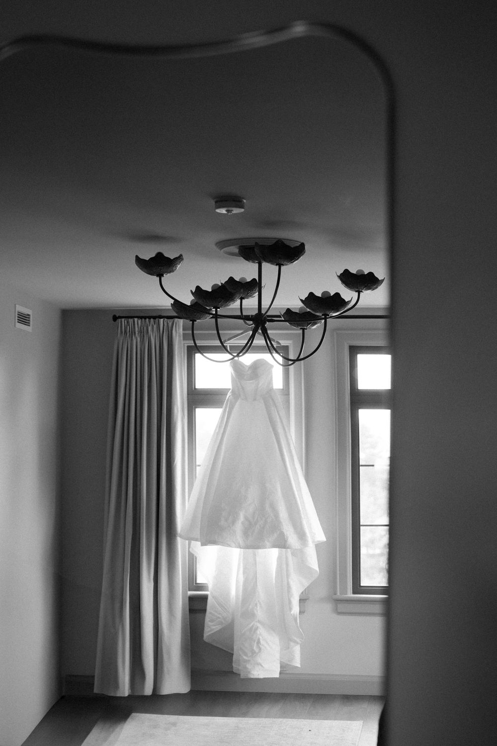 Black and white photo of an elegant white wedding dress hanging from a chandelier in a sunlit room.