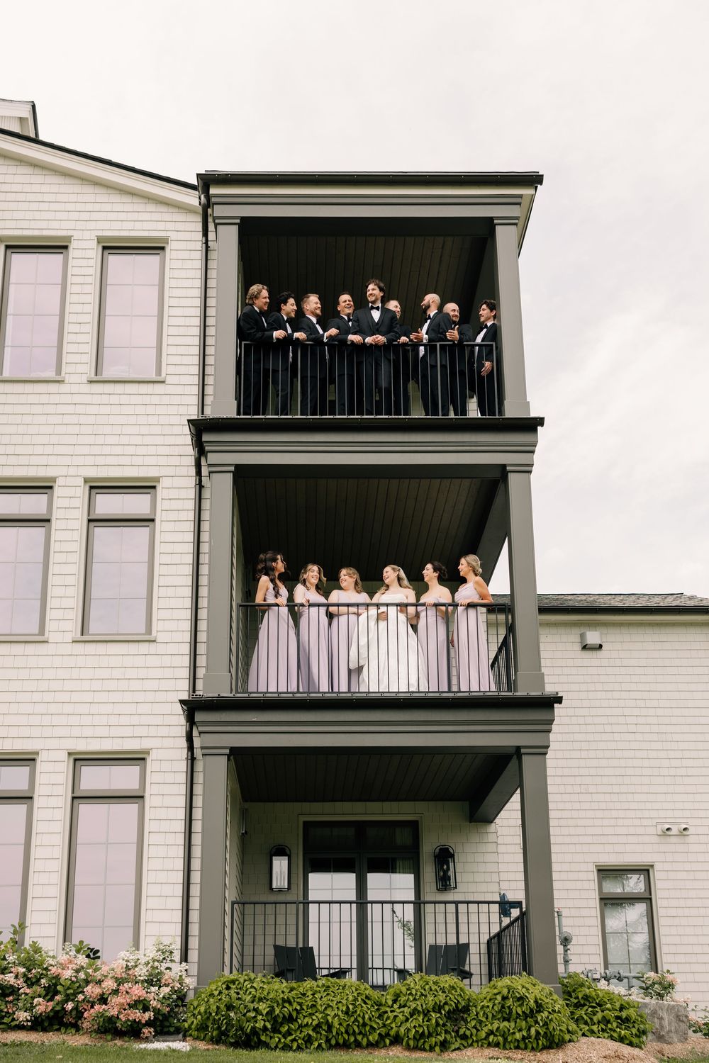 Wedding party posed on two balconies of a multi-story building, groomsmen on top and bridesmaids in blush gowns below.