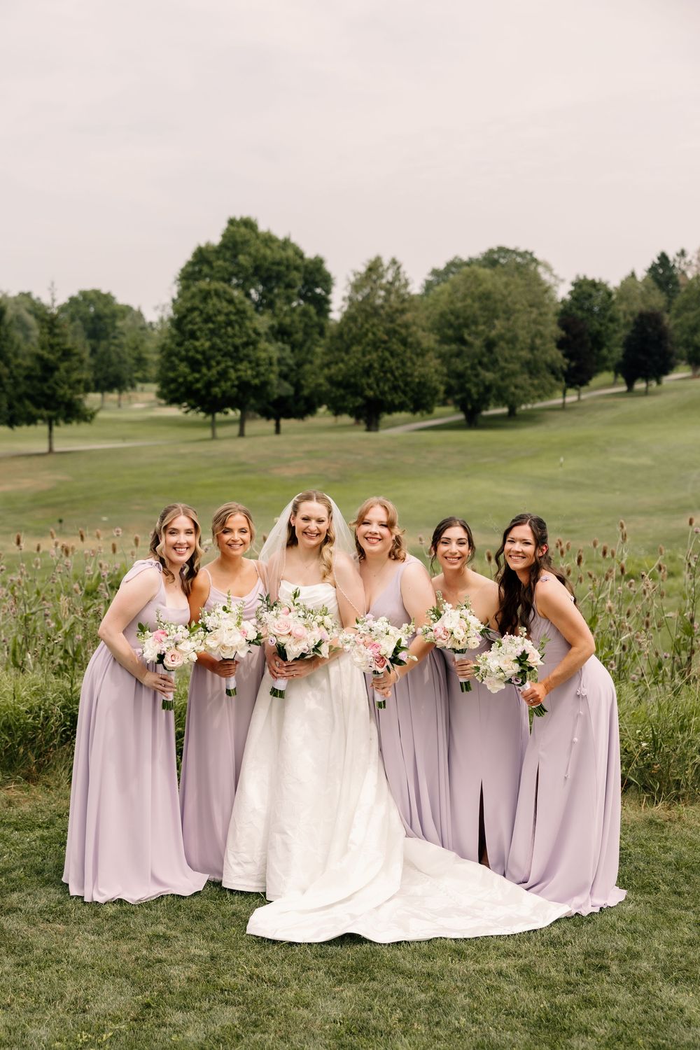 Bride in white gown with five bridesmaids in lavender dresses holding bouquets on a lush green golf course.