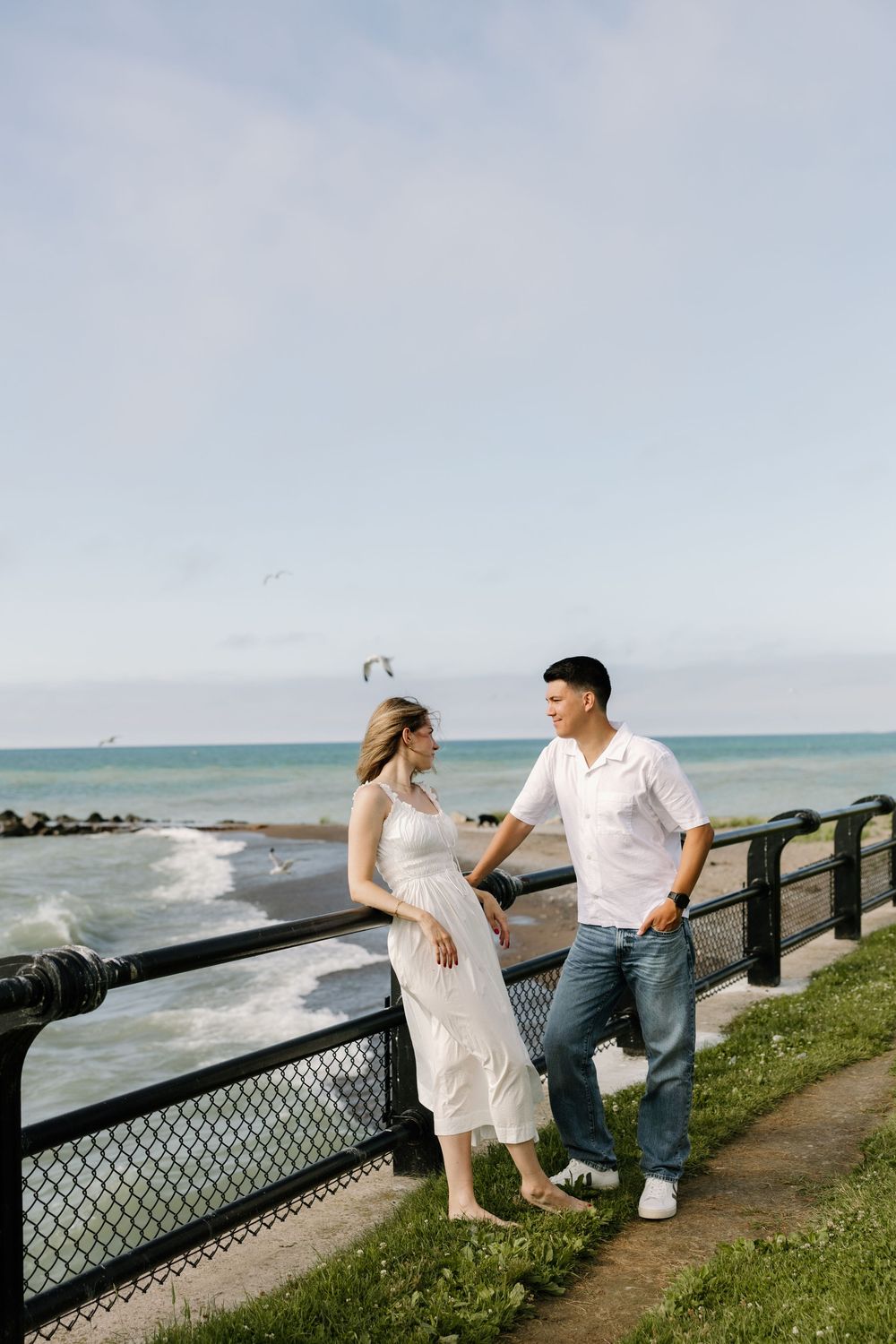 Couple in white outfits share a romantic moment by ocean railing on a breezy coastal walkway.
