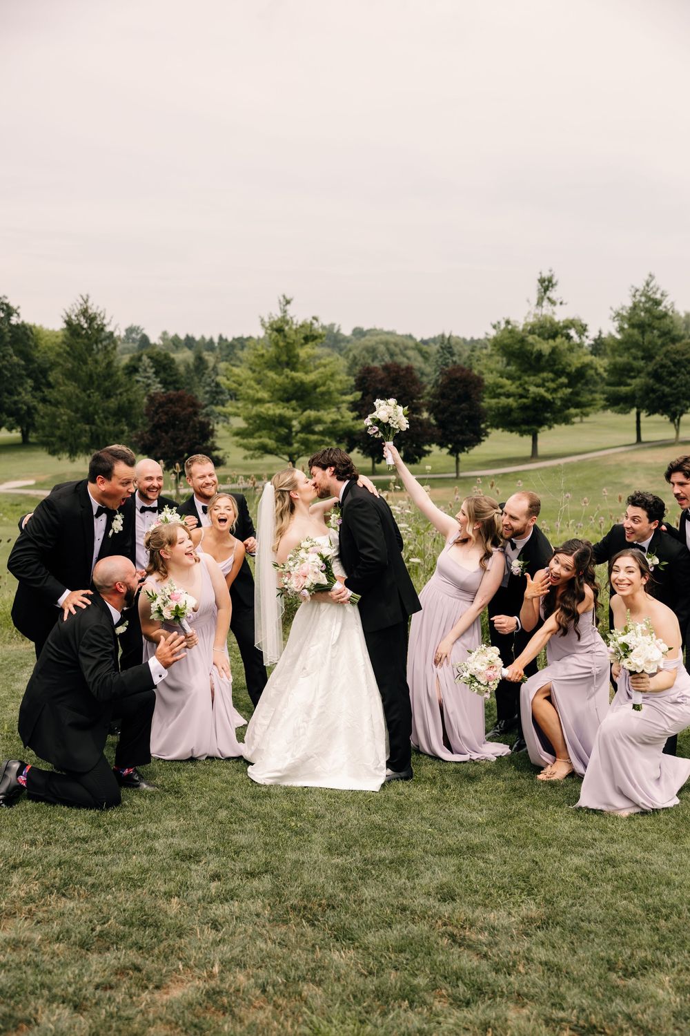 Wedding party celebrates as bride and groom kiss outdoors, bridesmaids in lavender gowns and groomsmen in tuxedos cheering.