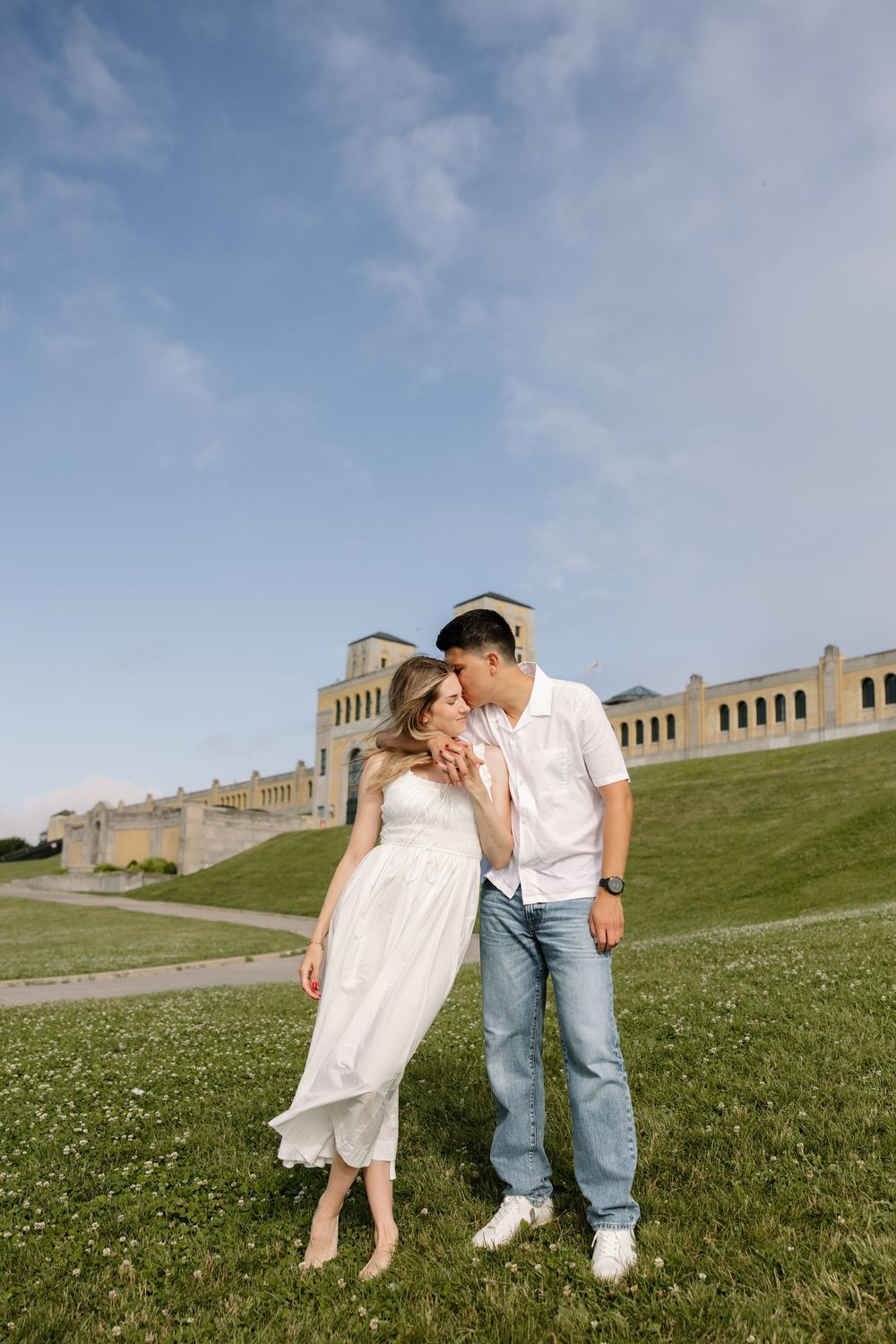 Couple embraces on green lawn with a historic stone building backdrop under blue sky.