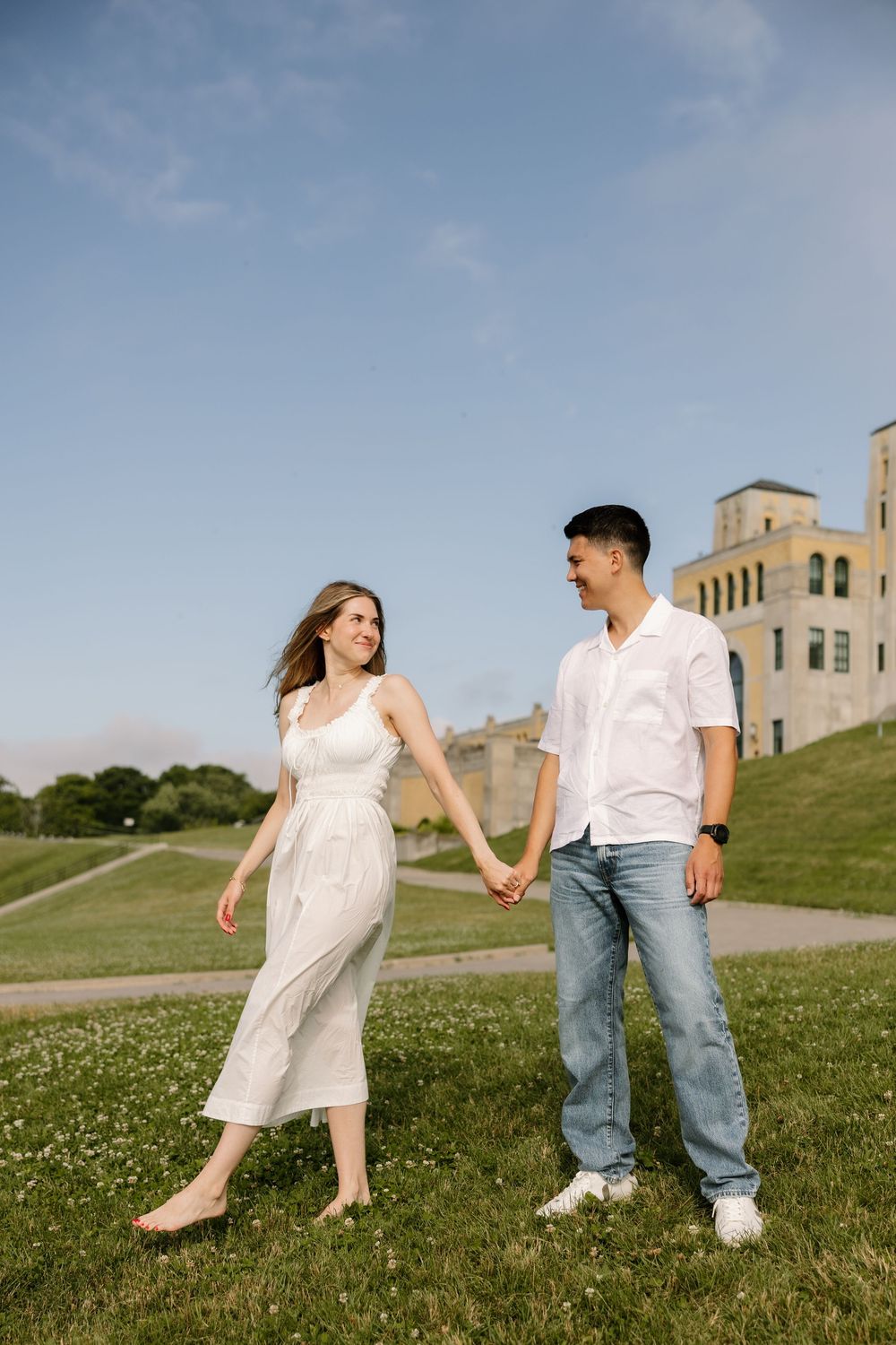 Happy couple holding hands and walking on a grassy hill with a historic stone building behind them.