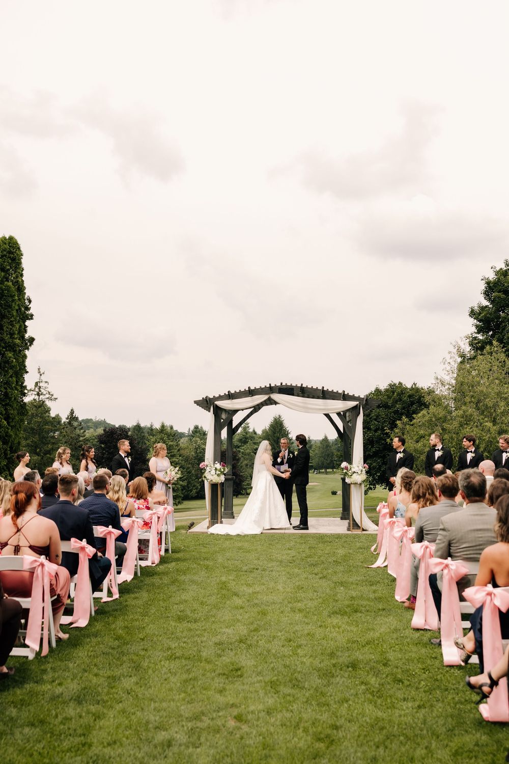 Outdoor wedding ceremony on green lawn with pink ribbon chair decorations leading to a wooden arch where bride and groom stand.