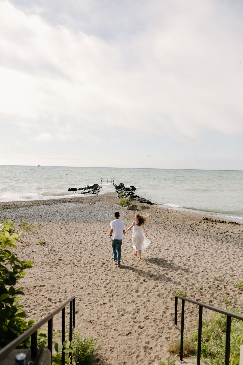 Couple walks hand in hand toward a rocky pier on a sandy beach under a cloudy sky.