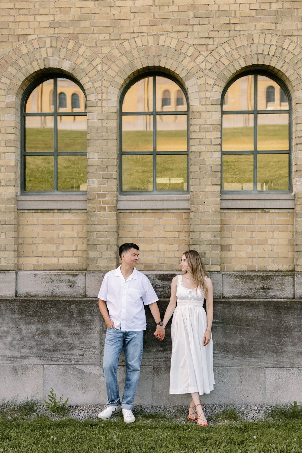 Couple holding hands in front of arched windows on a brick building exterior during an engagement photo session.