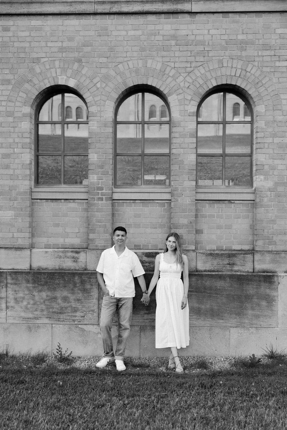 A couple holds hands in front of an arched brick building in this classic black and white engagement photo.
