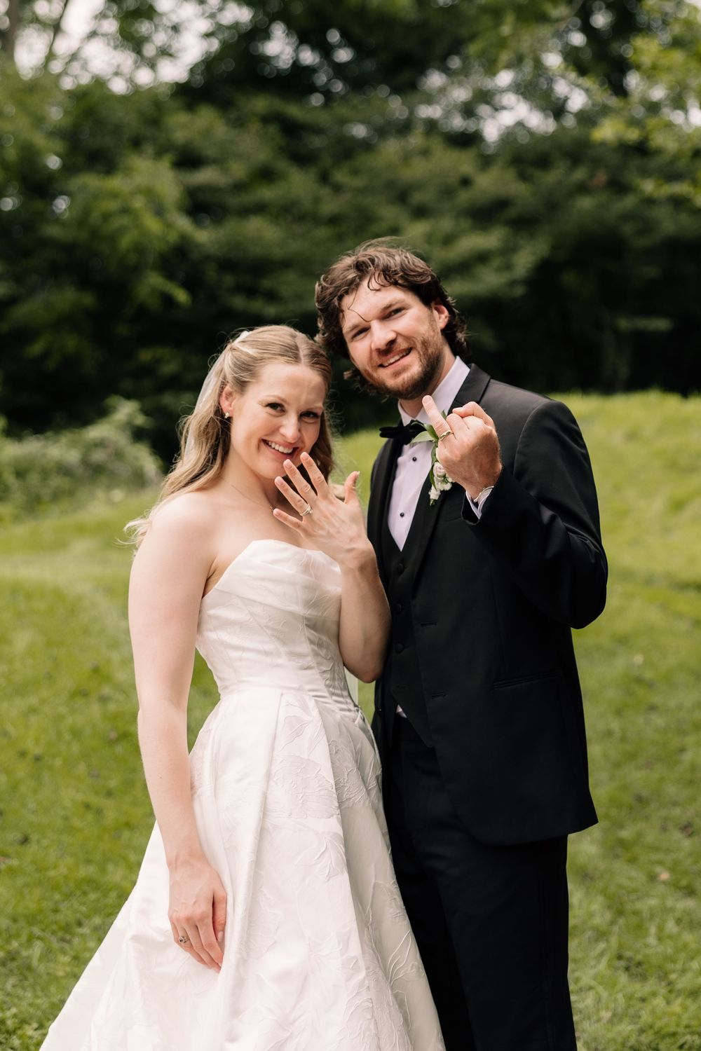 Bride and groom smiling outdoors showing wedding rings, bride in white gown, groom in black tuxedo with boutonniere.