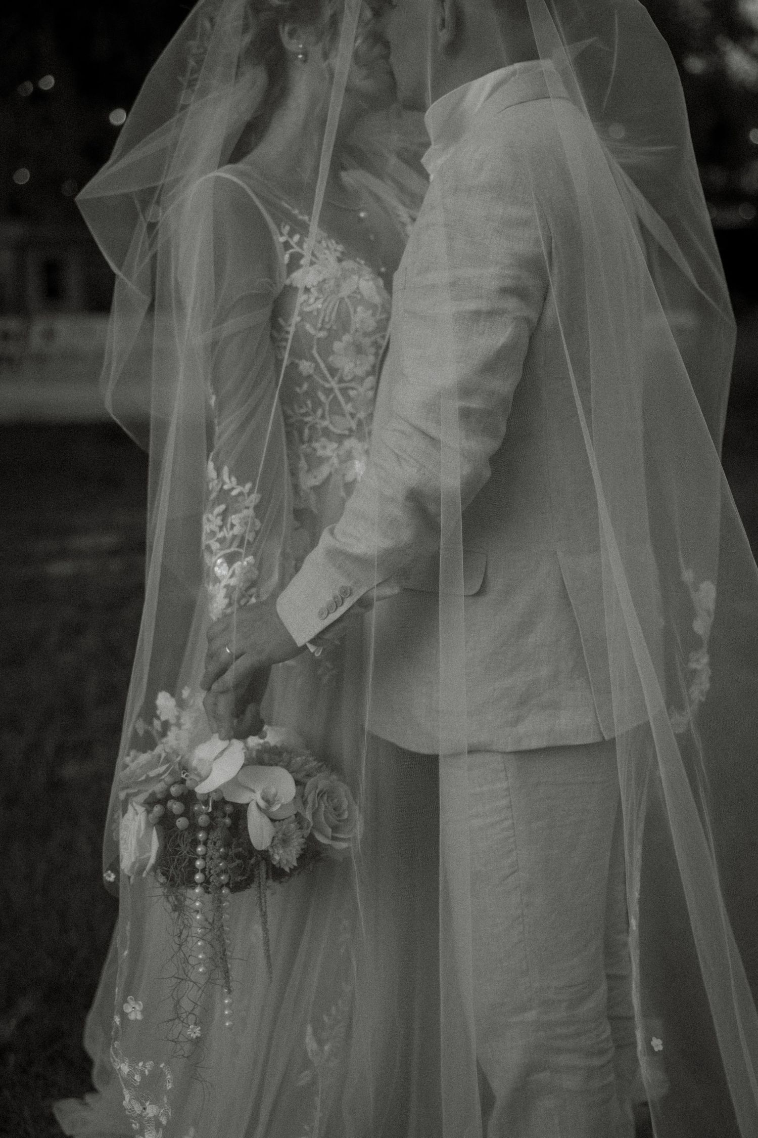 Black and white photo of bride and groom under veil, bride holding floral bouquet with pearls.