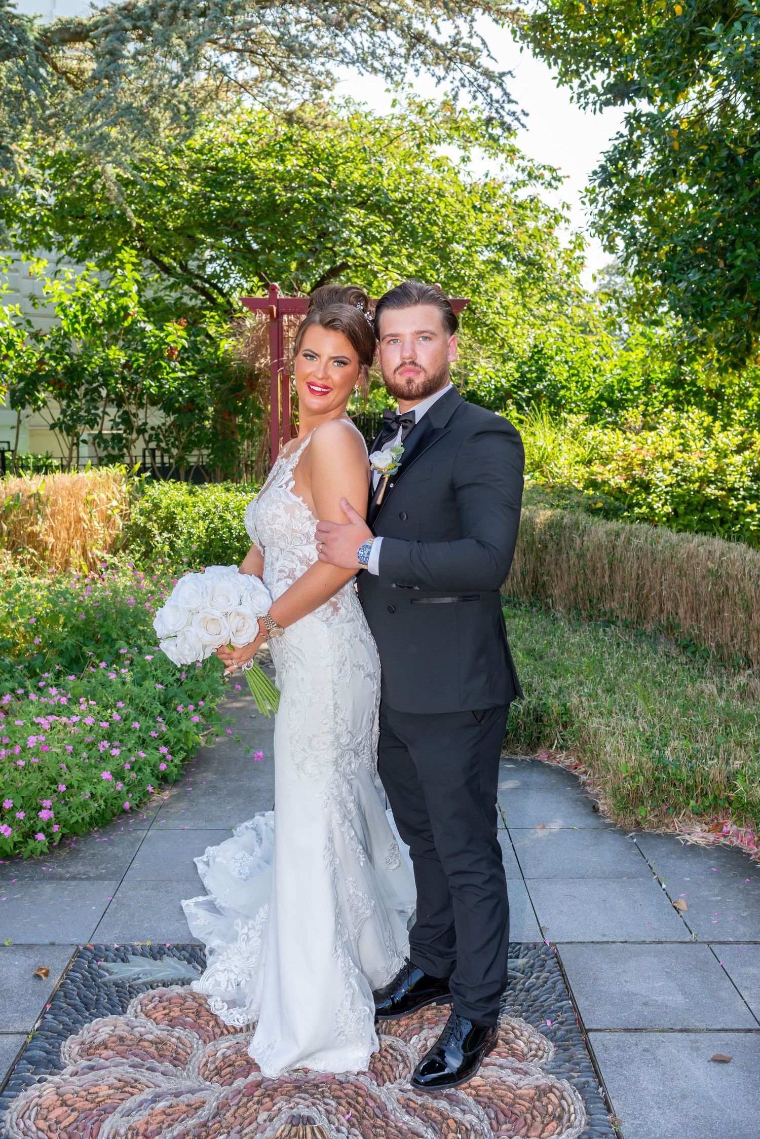 Bride in white lace gown holding bouquet poses with groom in black tuxedo in a lush garden setting.