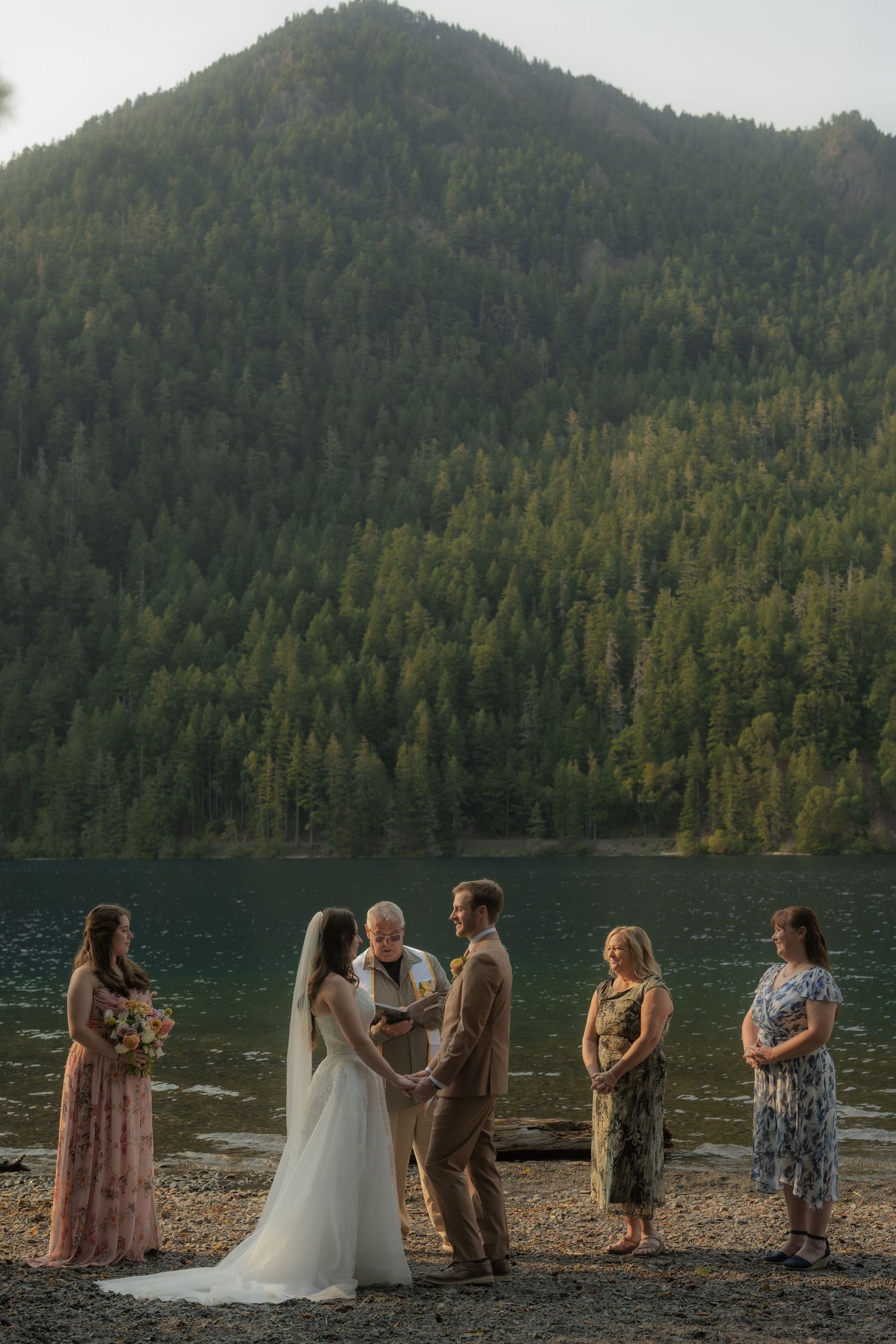 Outdoor lakeside wedding ceremony with mountain backdrop, couple exchanging vows with officiant and guests.