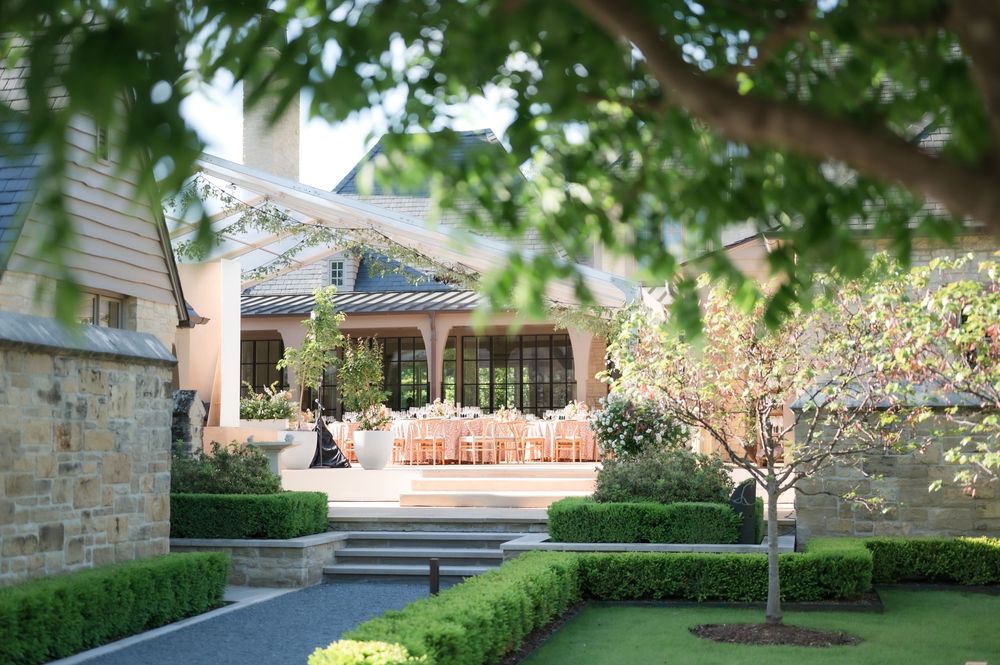 Elegant garden entrance with stone steps leading to covered porch through manicured boxwood hedges and blooming trees.