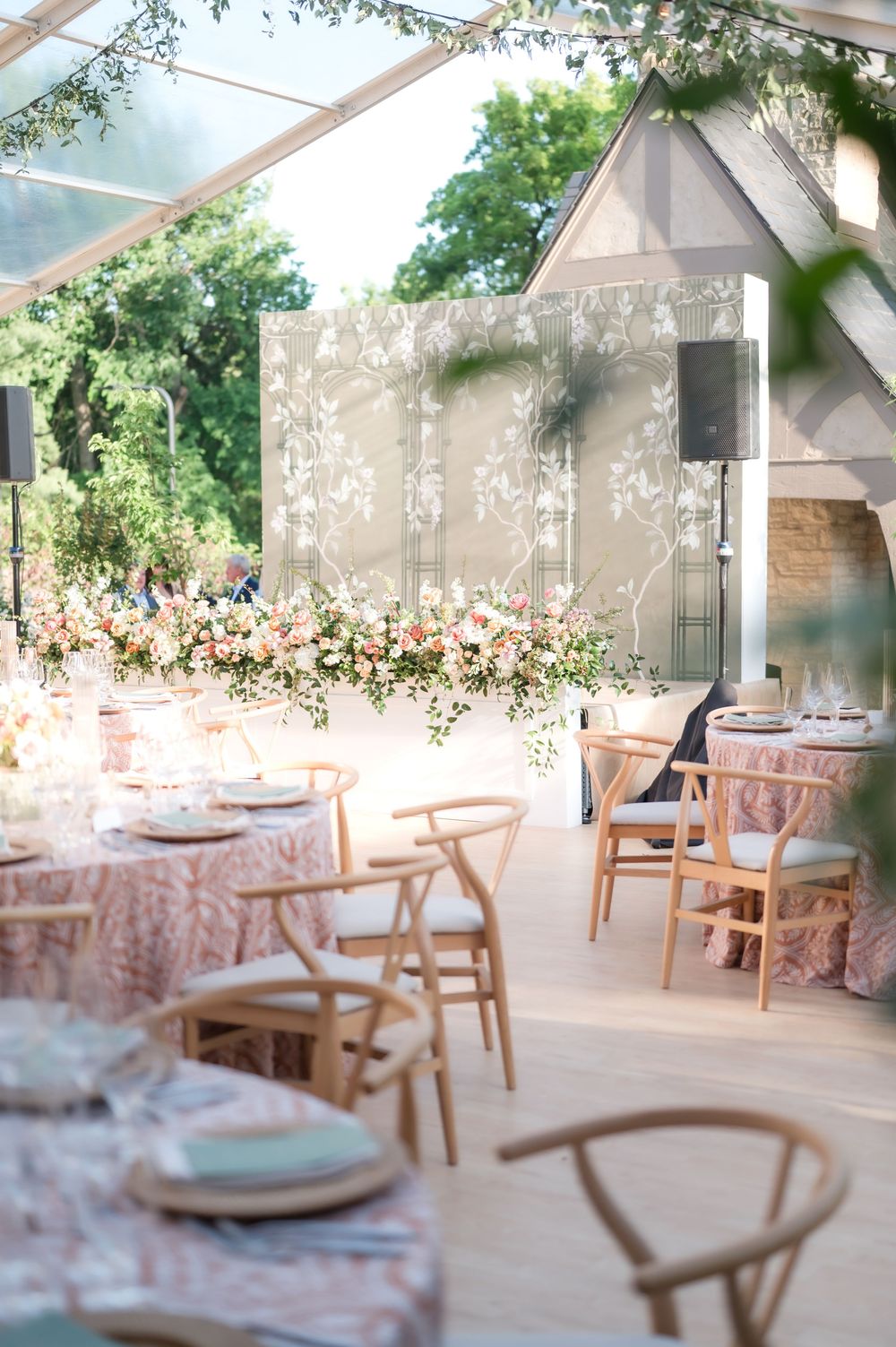 Elegant wedding reception setup with soft pink tablecloths, wooden chairs and floral arrangements under a glass-covered venue.