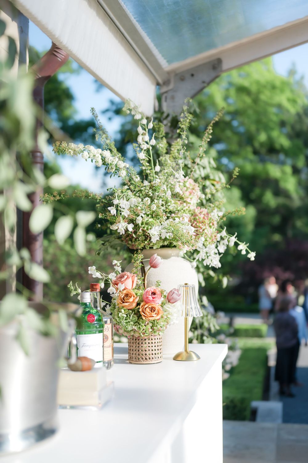 Delicate floral centerpiece with white wildflowers and pink roses in a textured vase on outdoor wedding bar counter.