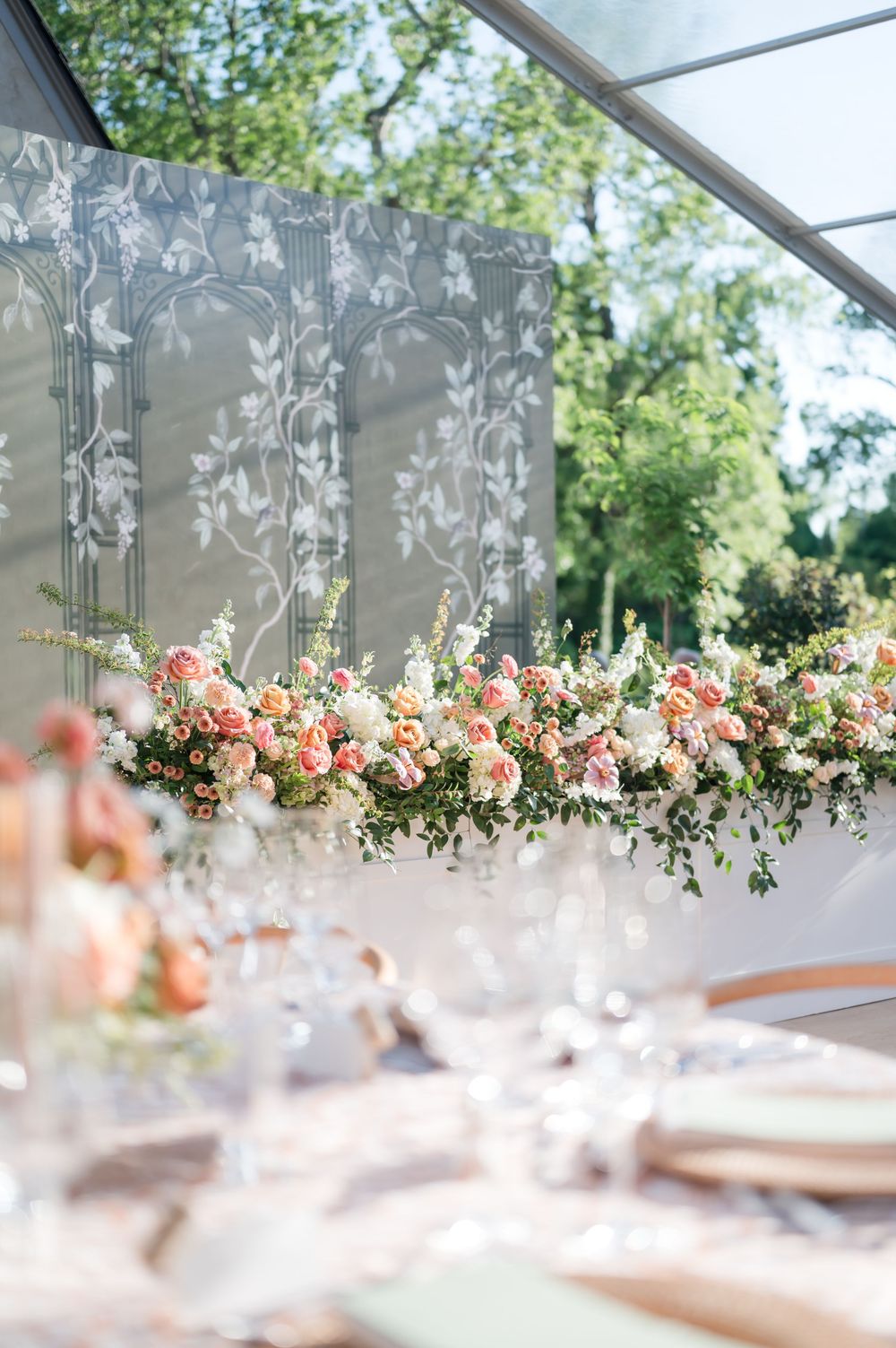 Elegant floral arrangement with pink and peach roses adorns a wedding reception table under a draped tent canopy.