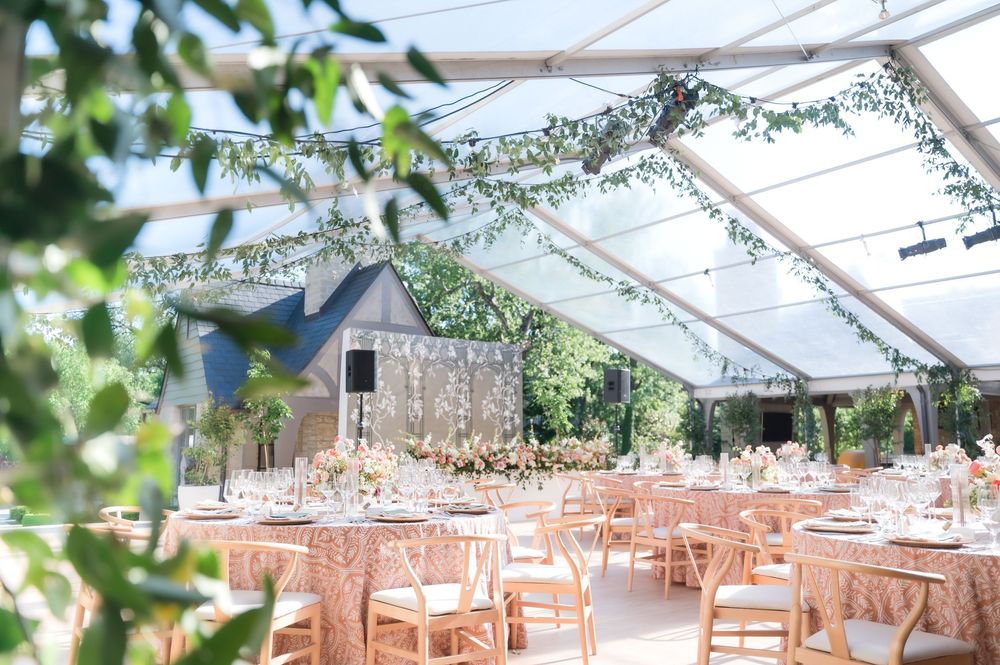 A clear-top wedding tent decorated with greenery and filled with peach-colored tables and chairs for an elegant reception.