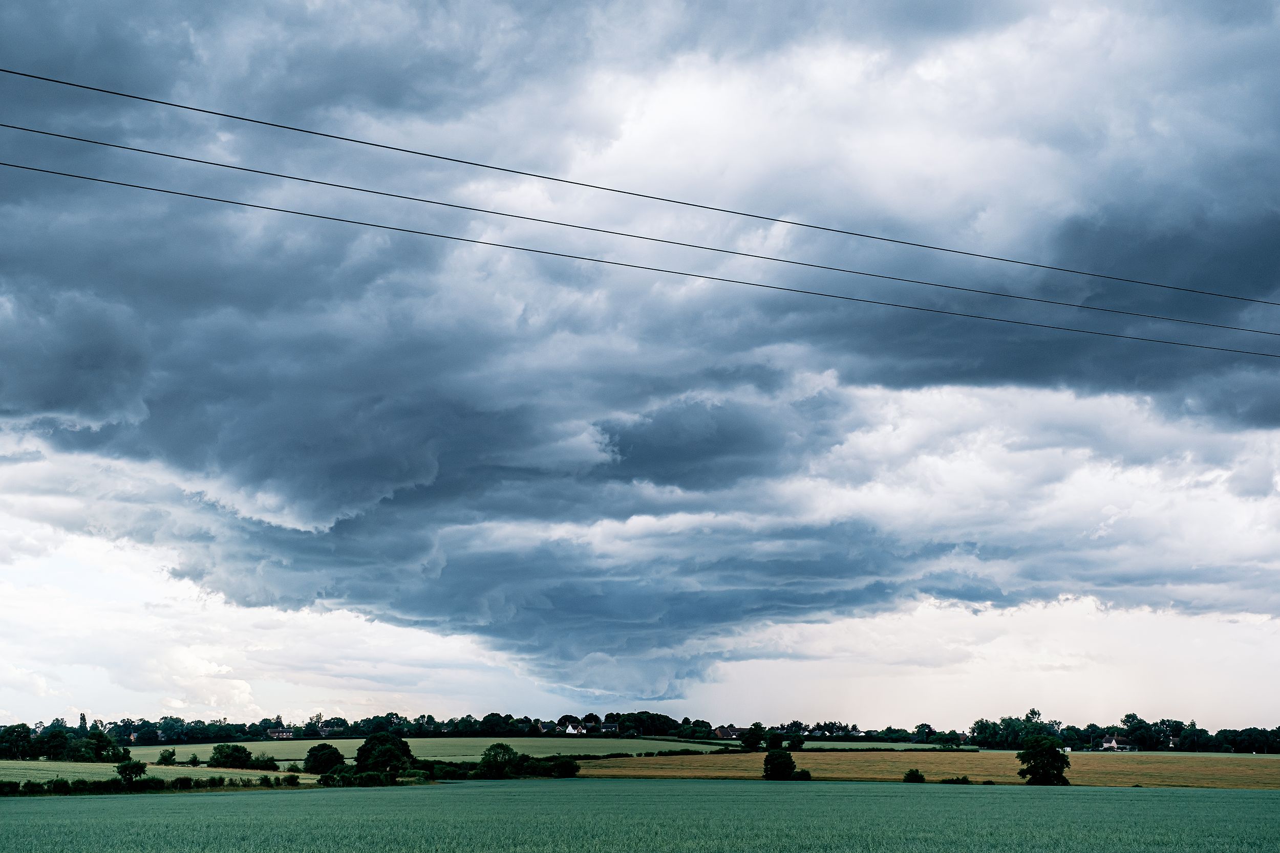 Dramatic storm clouds loom over lush green farmland with power lines stretching across the moody sky.