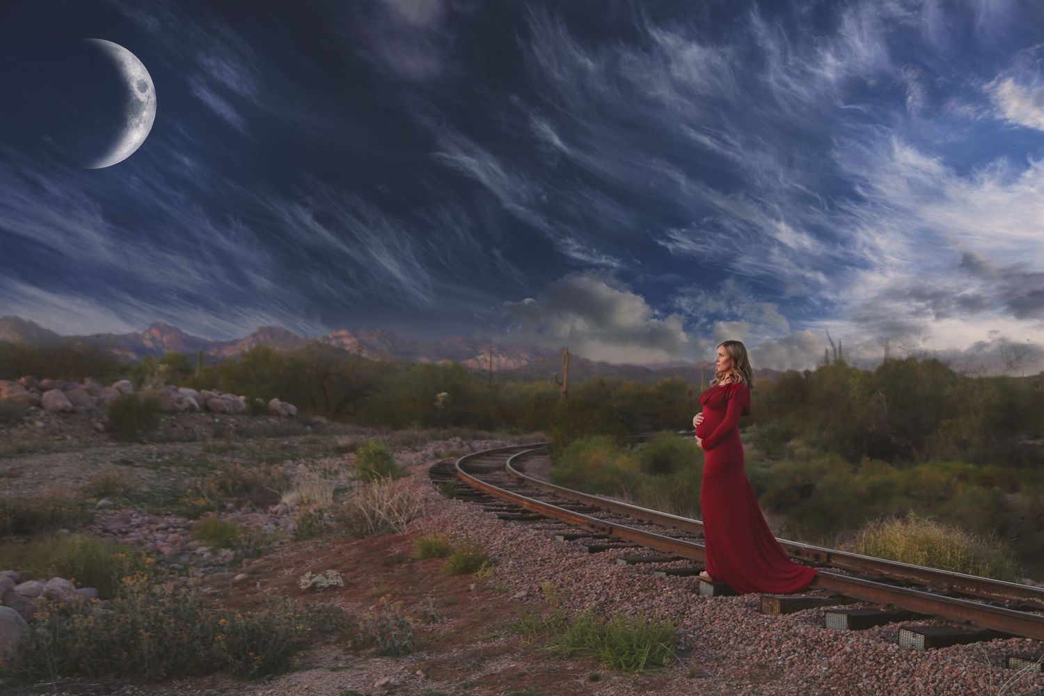 Woman in a flowing red gown stands beside railroad tracks in a desert landscape under a dramatic night sky with a crescent moon.