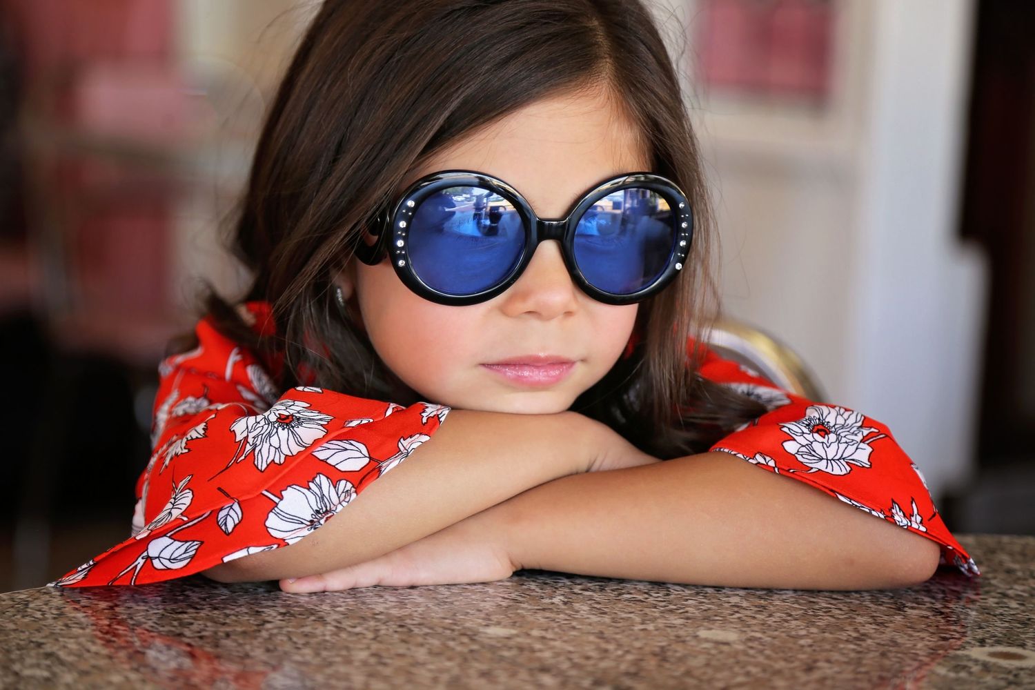 Young girl in blue round sunglasses and a red floral top rests her arms on a granite countertop.