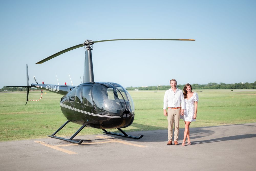 A couple stands beside a sleek black helicopter on an airfield tarmac.