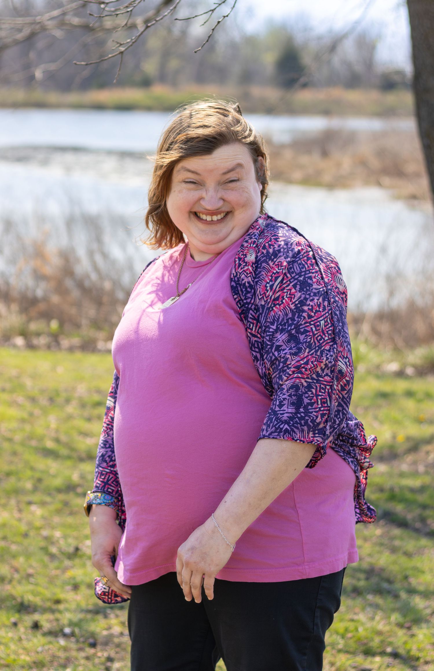 Smiling woman in pink top and floral cardigan posing outdoors near a lake on a sunny spring day.