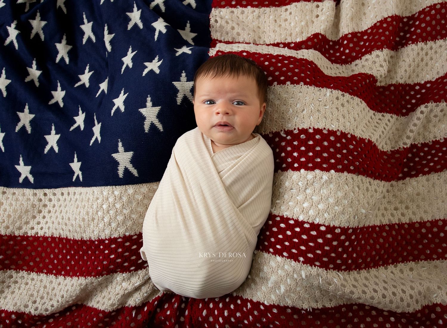 Newborn baby swaddled in white wrap posed on an American flag blanket with red stripes and white stars.