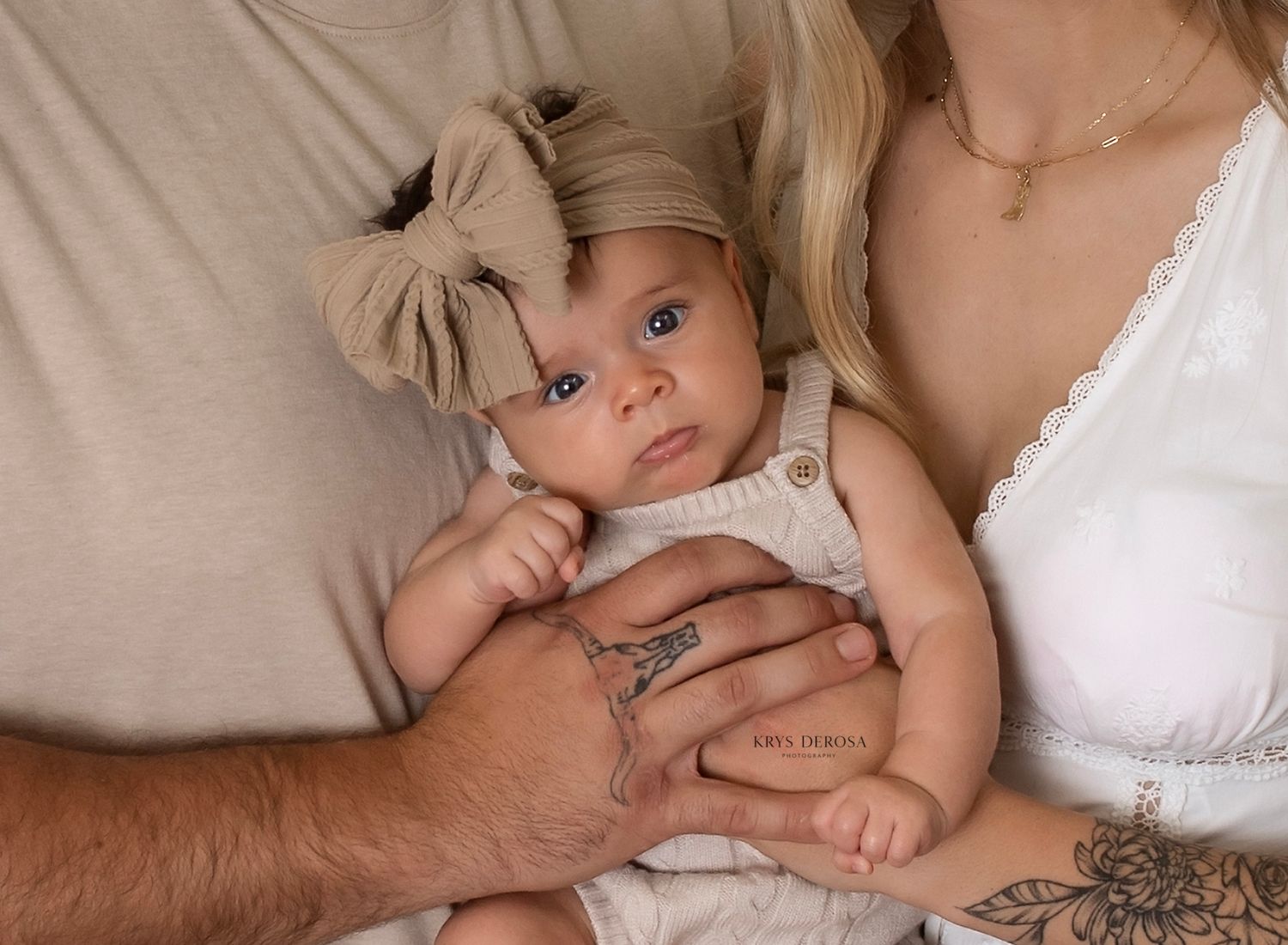 Newborn baby in beige bow headband cradled by parents with tattoos and gold jewelry.