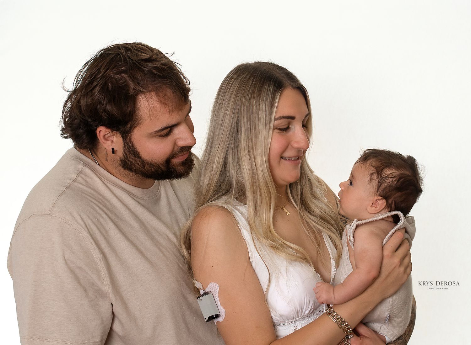 A young couple smiles lovingly at their newborn baby during a professional studio photography session on white background.