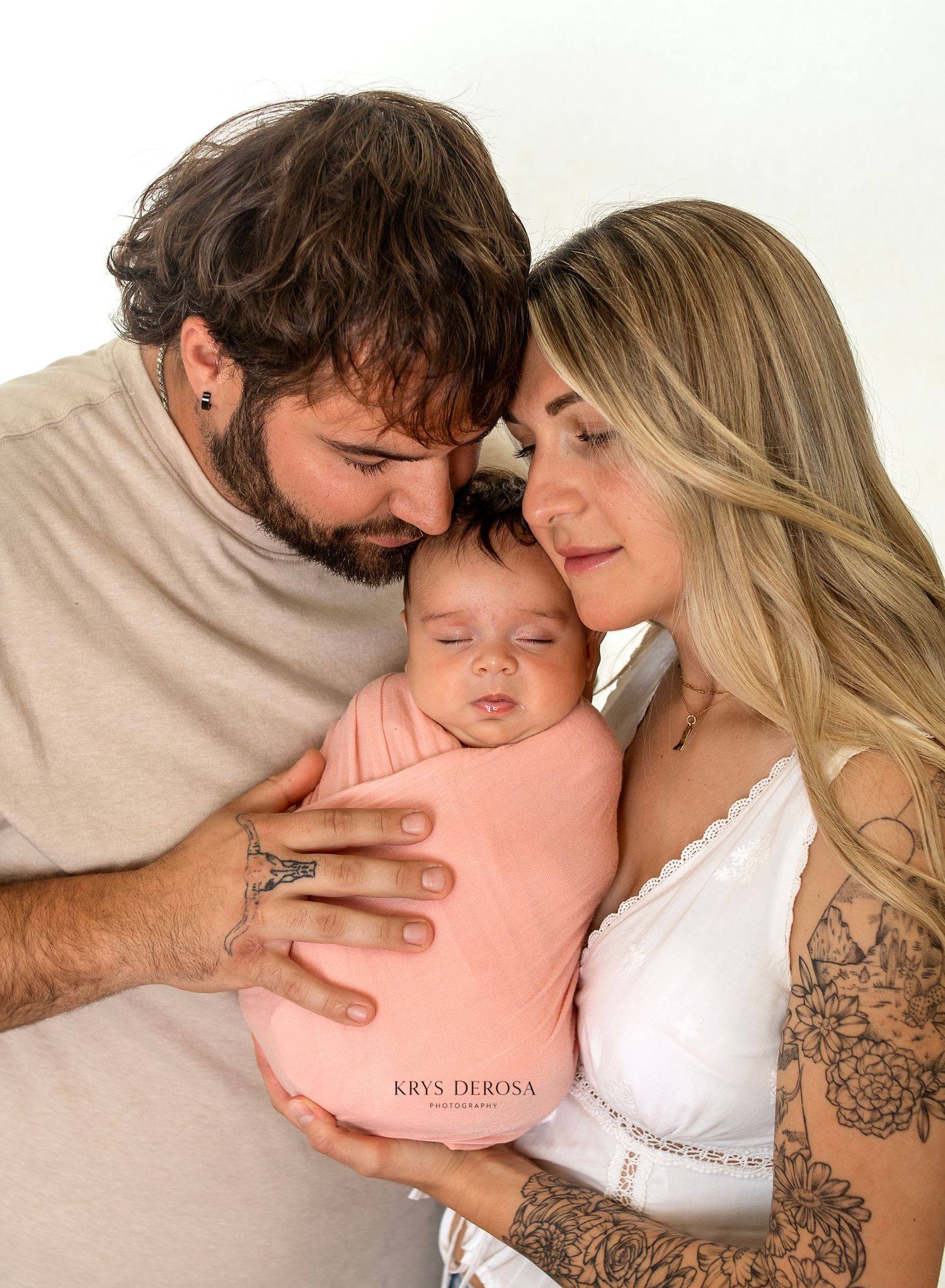 New parents tenderly kiss their sleeping newborn wrapped in a pink swaddle during a professional newborn photo session.