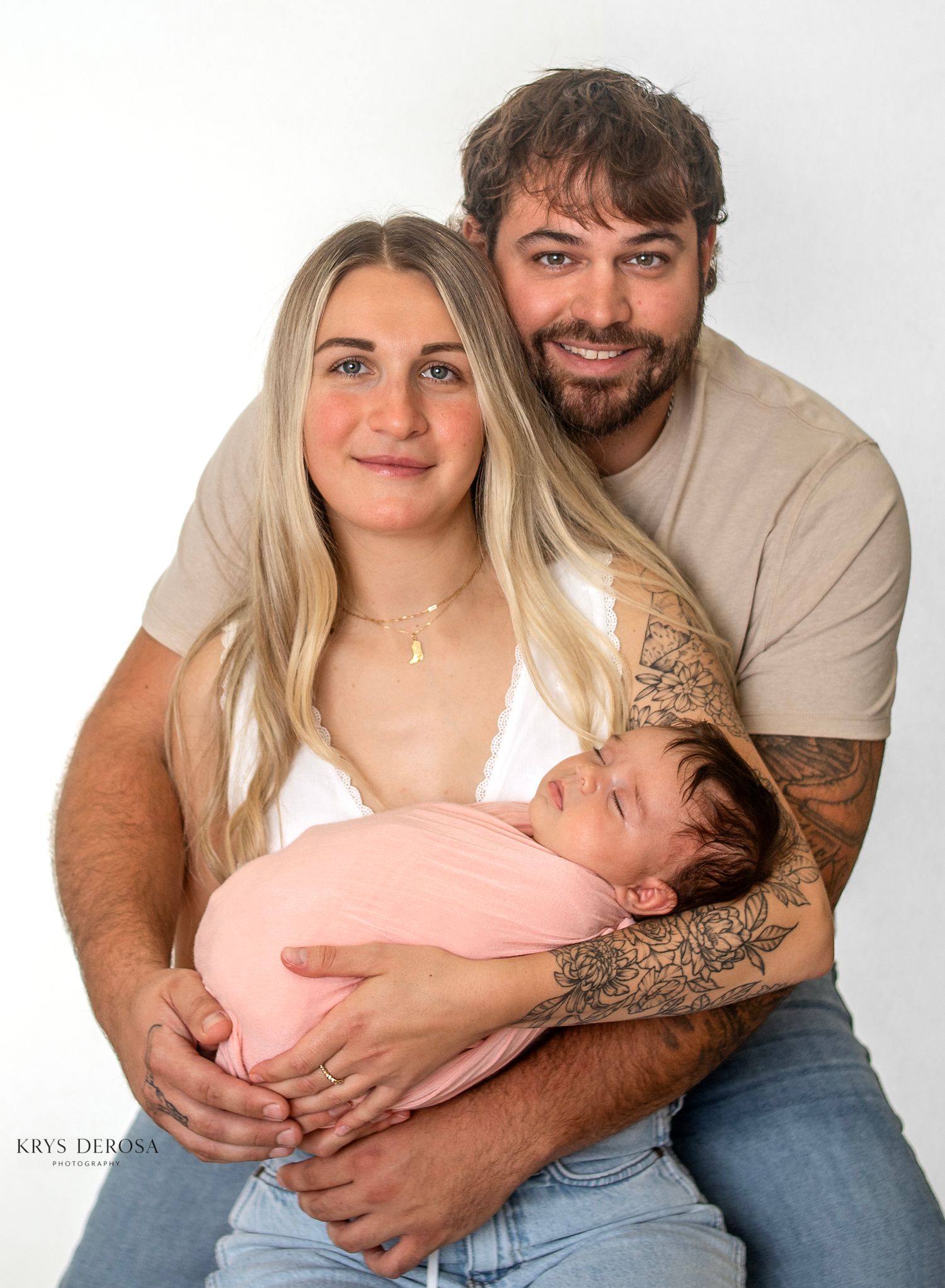 Young couple smiling and holding their newborn baby wrapped in pink during a professional studio portrait session.