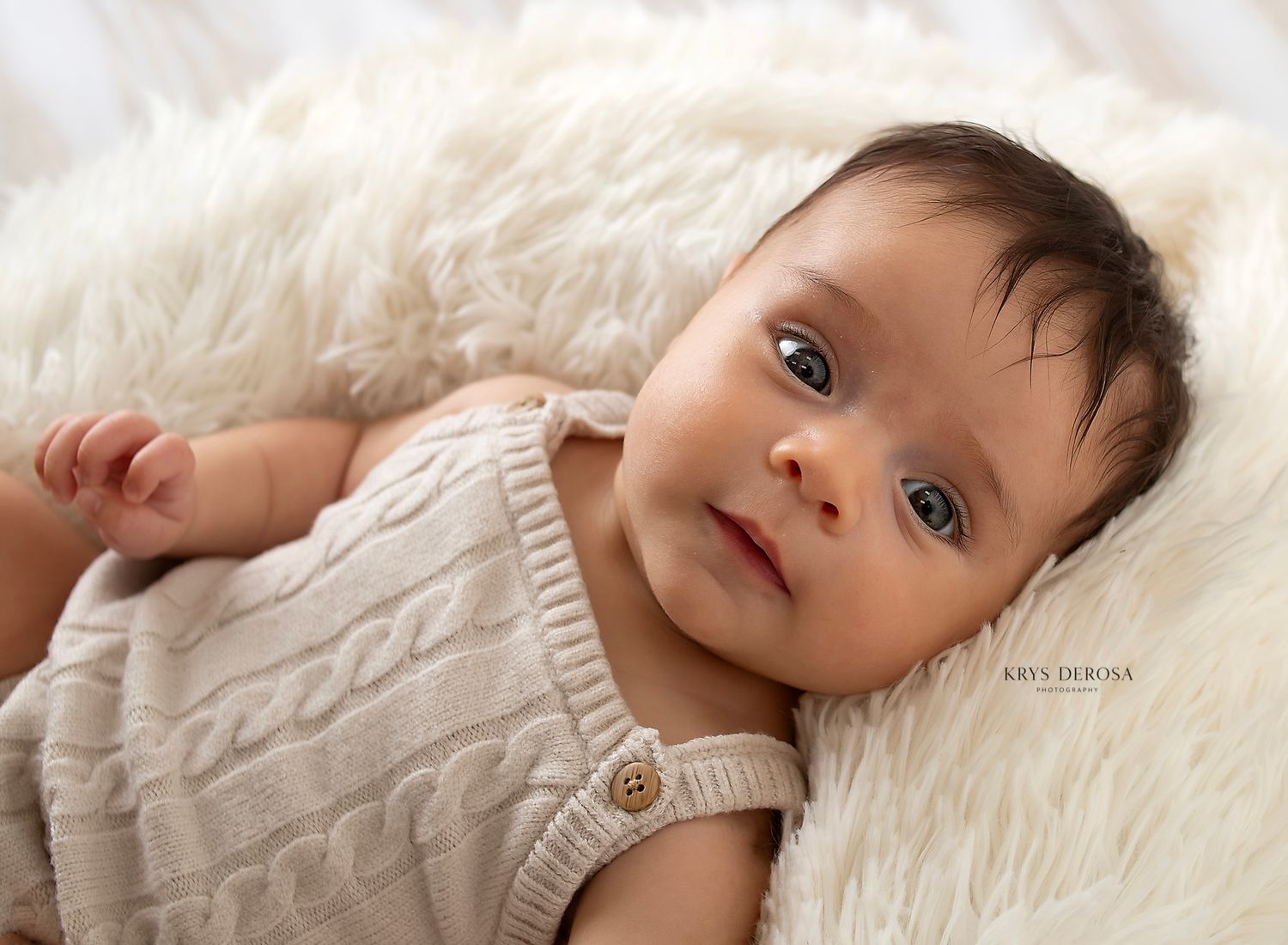 A serene baby in a cream knit romper lies on a fluffy white blanket, gazing curiously with bright eyes.