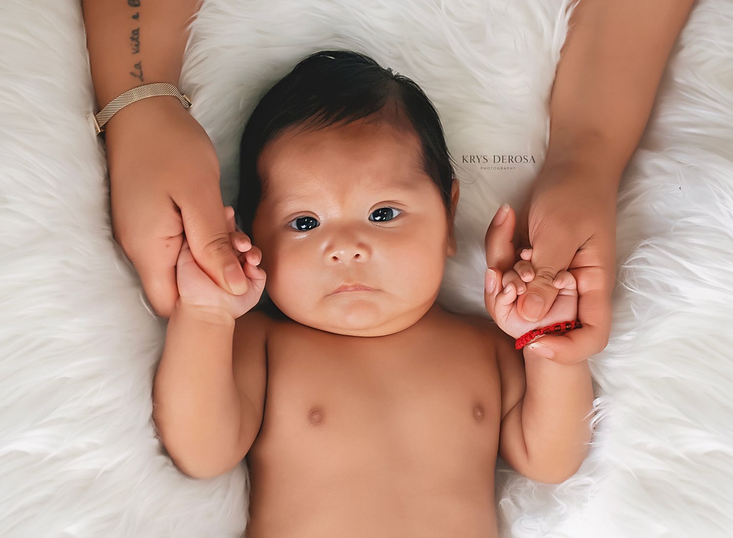 Adorable alert newborn baby lies on white fur rug while parents gently hold tiny hands in a professional photo.