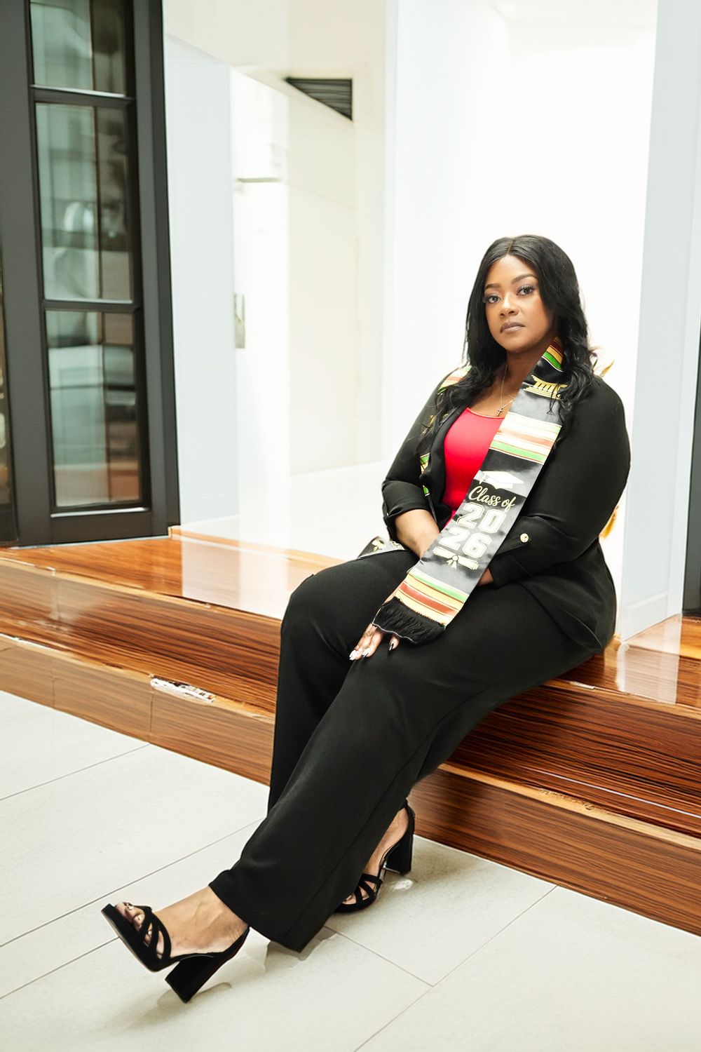 Woman in black suit and colorful graduation sash sits on wooden steps in modern building interior.