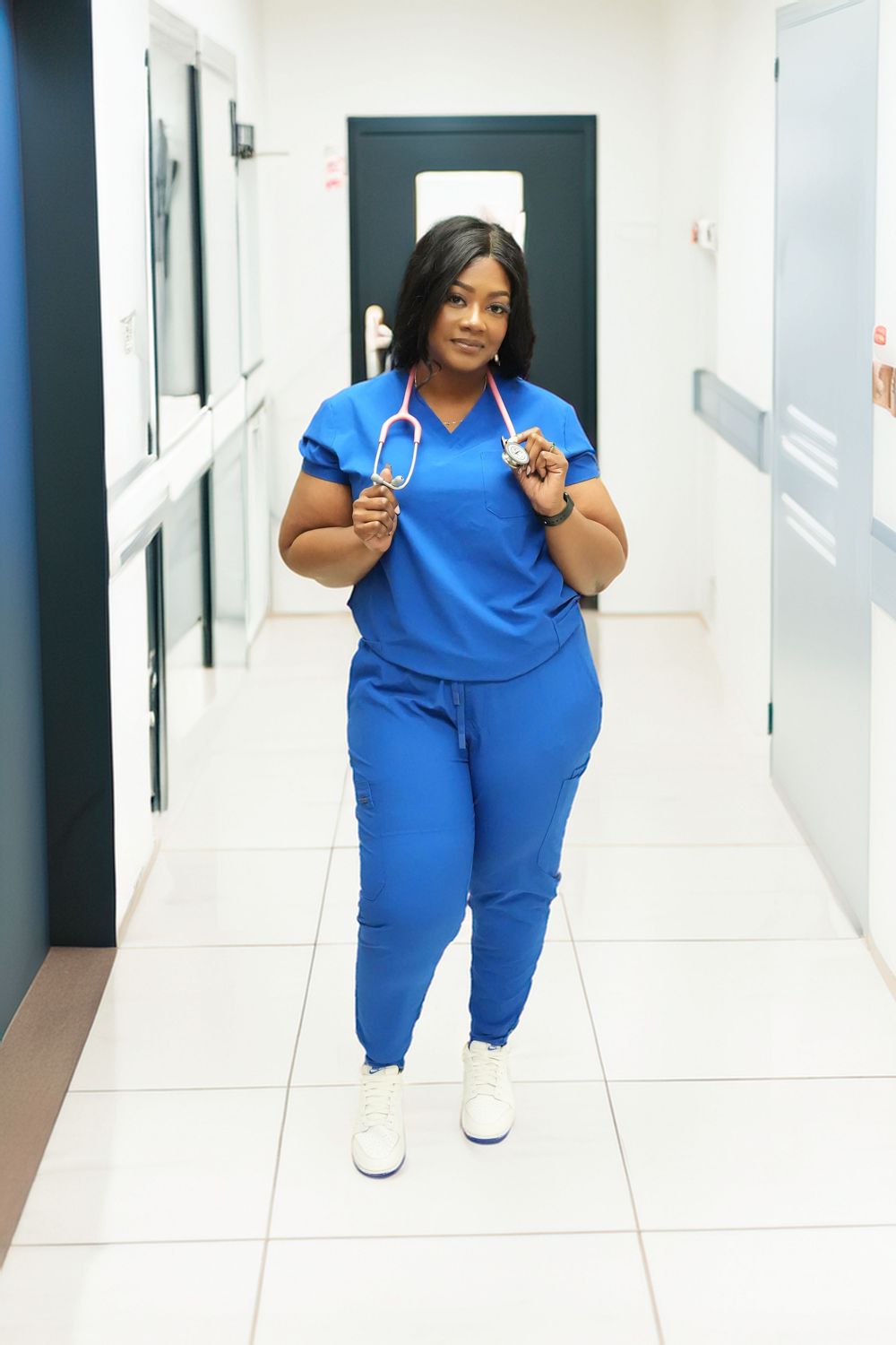 A female nurse in blue scrubs holds a stethoscope while walking confidently down a hospital corridor.