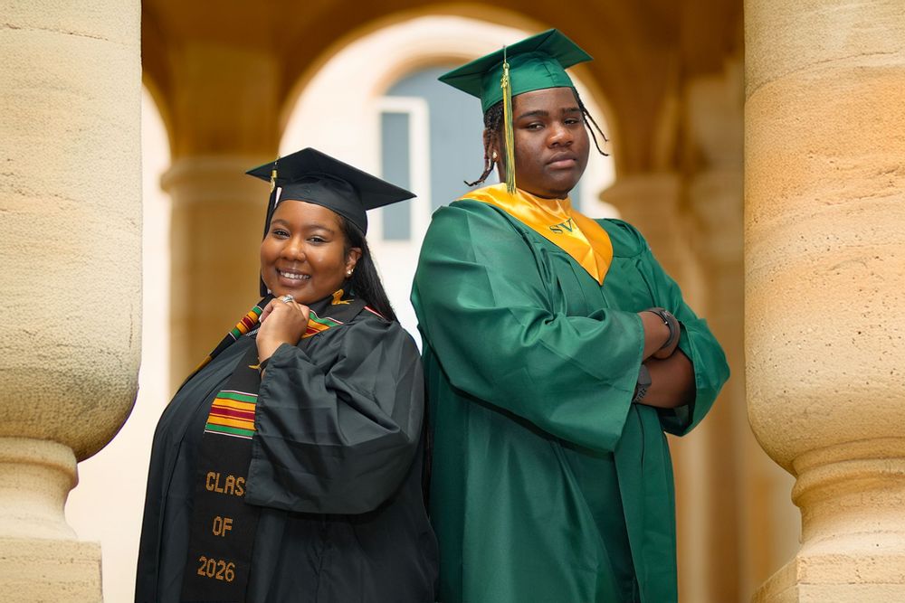 Two graduates in caps and gowns, one in black and one in green, pose proudly between stone columns.