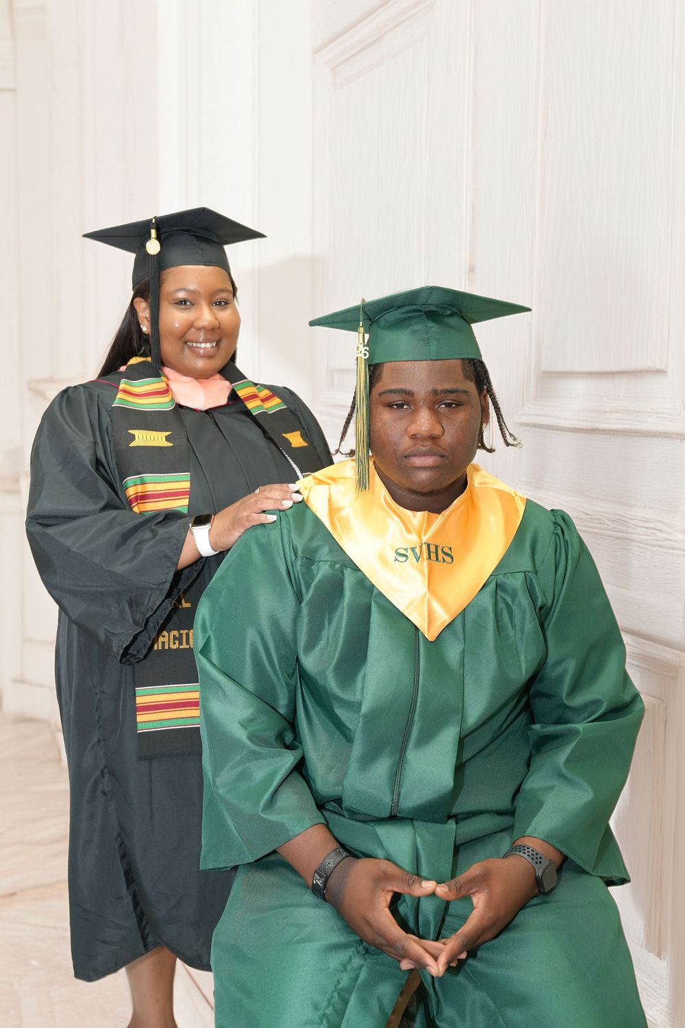Two graduates in green caps and gowns pose together, with one placing a hand on the other's shoulder, in a bright white room.