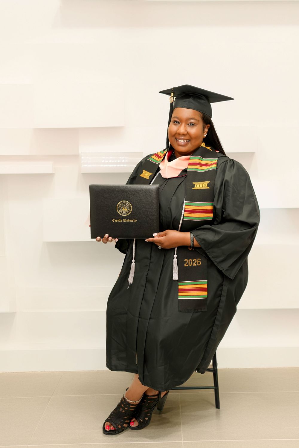 Graduate in black cap and gown holds diploma, wearing kente stole, posing proudly for graduation photo.